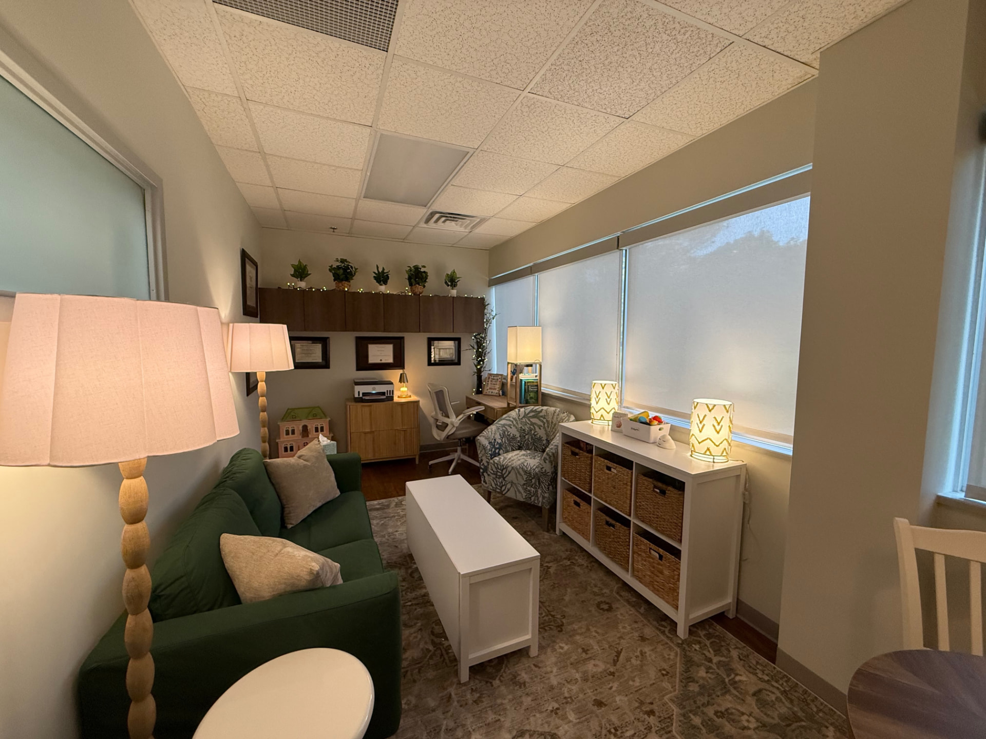 The same room from the doorway: a desk and chair, storage baskets, a wooden dollhouse on the floor, and bright windows.