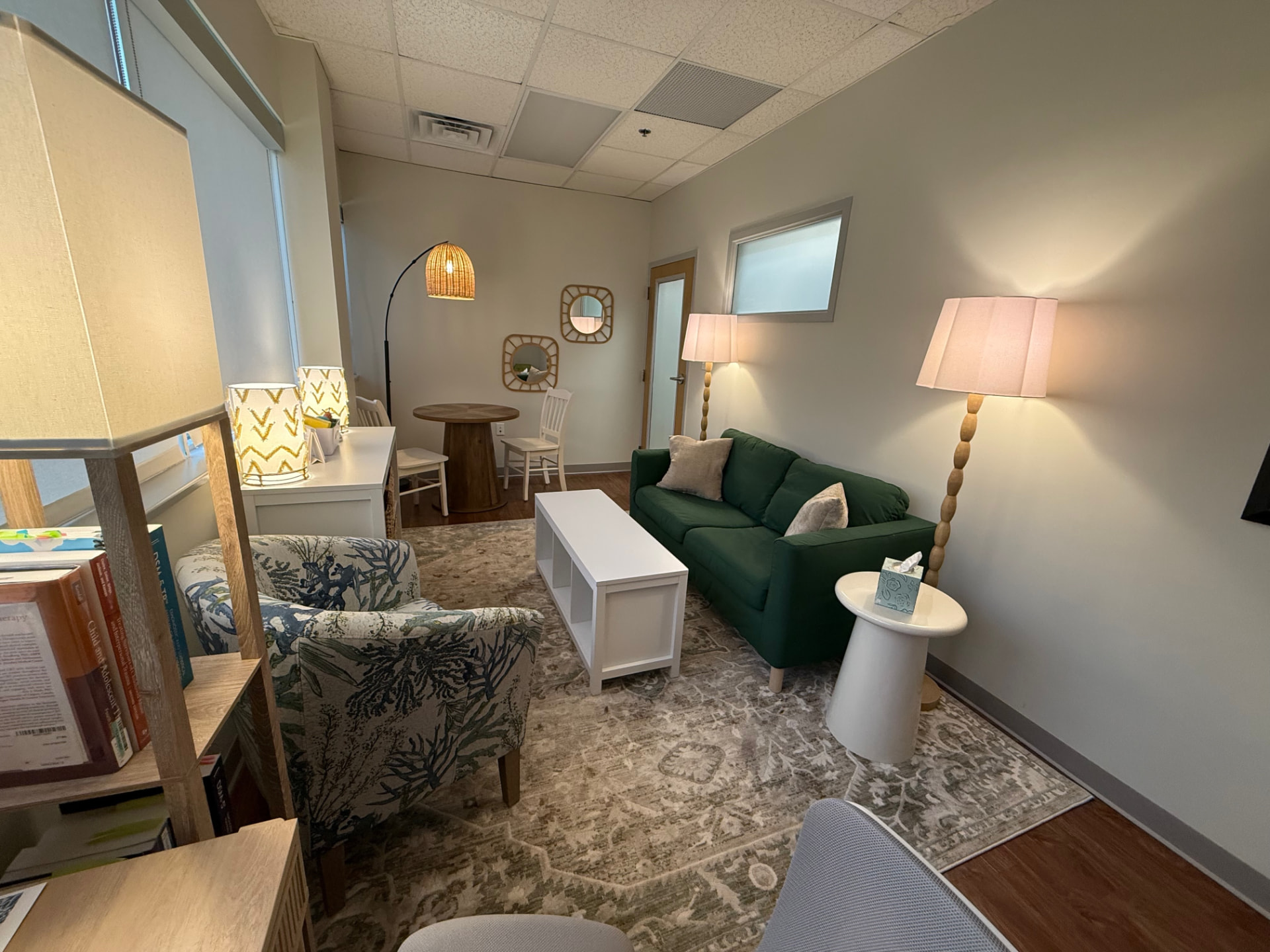 The therapy room: a green sofa, a patterned armchair, warm floor lamps, and a shelf of books.